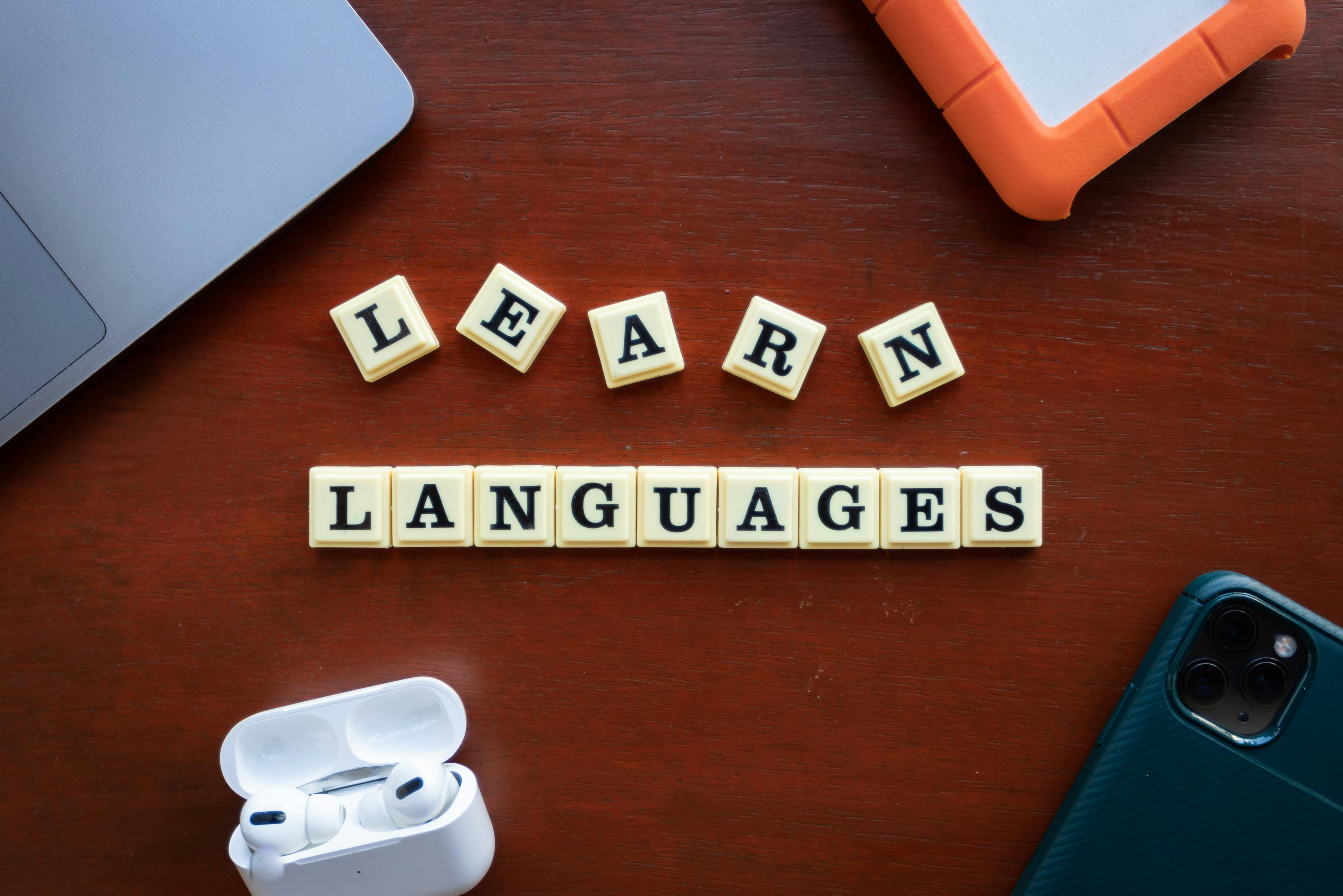 Scrabble kostičky se slovem „Learn languages“, doplněné o bezdrátová sluchátka a mobil, symbolizují moderní a motivující přístup k učení cizích jazyků.