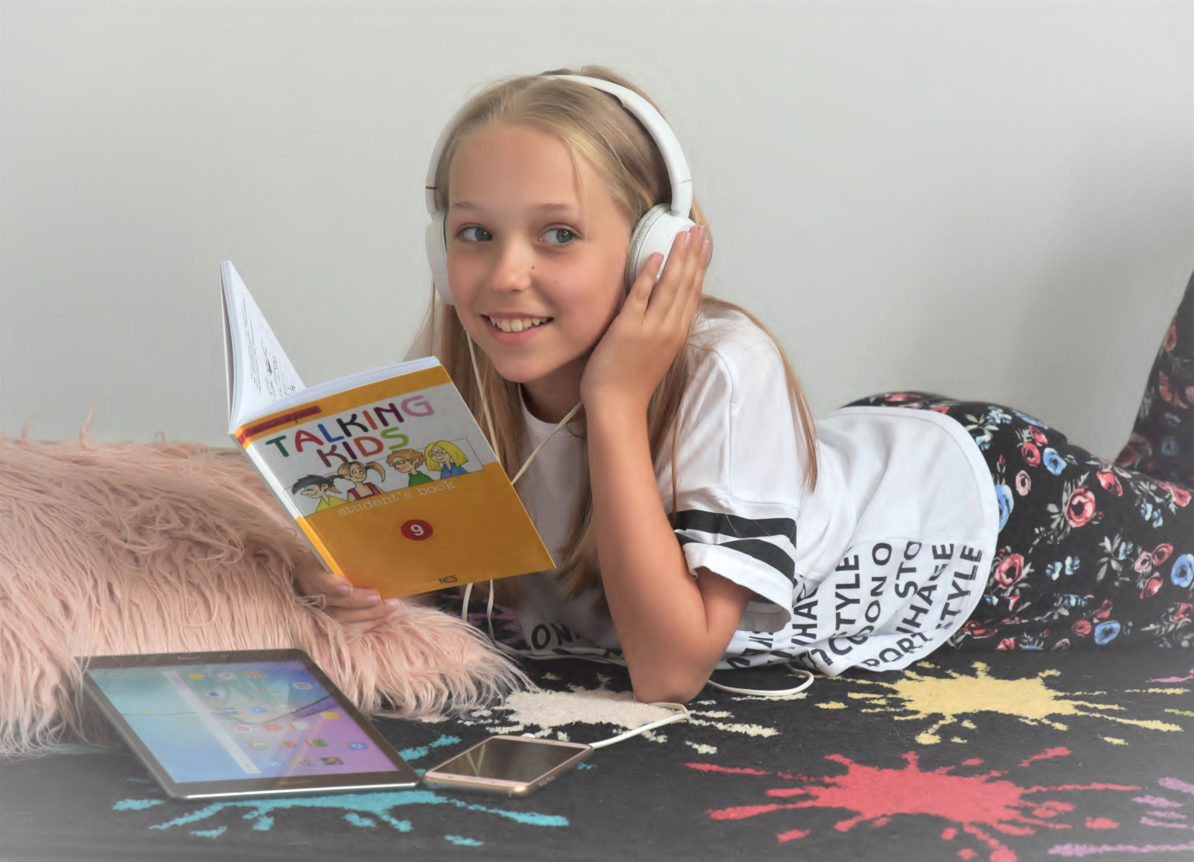 Dívka se sluchátky leží na posteli a učí se angličtinu z učebnice Talking Kids, moderní a zábavná forma výuky angličtiny pro děti z pohodlí domova.